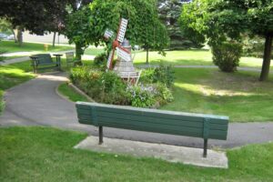 A peaceful park scene with green benches along a paved walking path, surrounding a small flower bed featuring a decorative windmill and lush greenery.