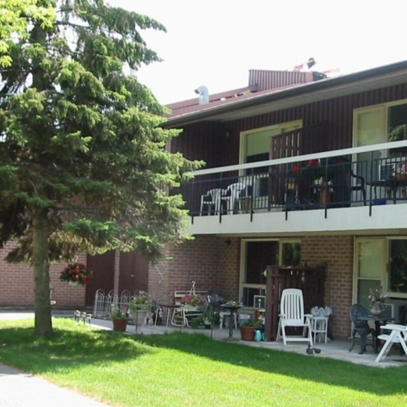 Aldersgate two story apartment building with balcony surrounded by green grass and mature pine tree
