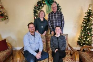 A group of four smiling people, two men and two women, pose in a living room decorated for Christmas. Two people are seated on a sofa with patterned cushions, and two people are standing behind them. A Christmas wreath hangs on the wall, and a decorated Christmas tree with a gold star on top stands to the right.